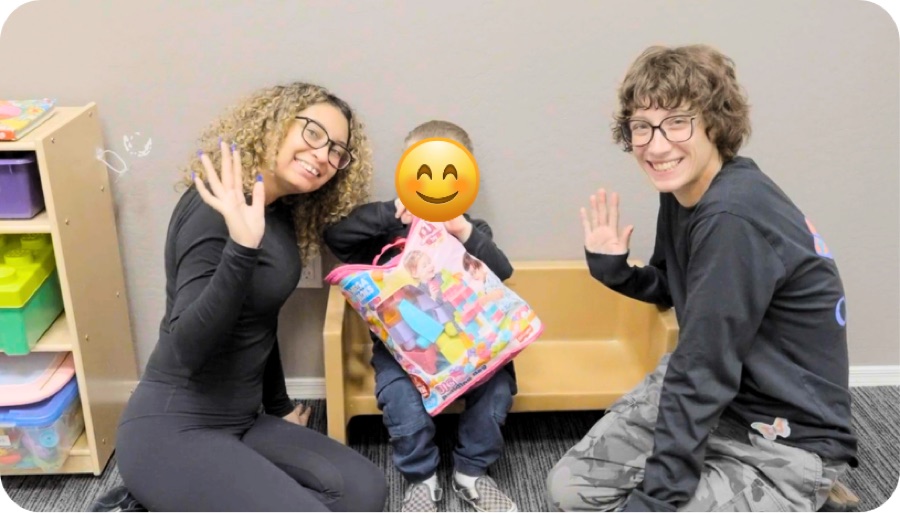 Two smiling team members waving beside a child holding a new toy